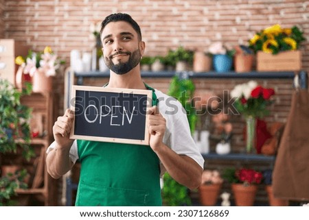 Hispanic young man working at florist holding open sign smiling looking to the side and staring away thinking. 