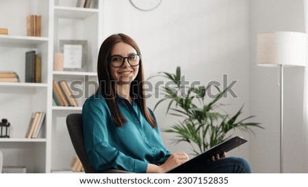Happy professional psychologist woman sitting in chair writing on clipboard, looking and smiling at camera, working in modern office. Psychotherapeutic services, mental health professional concept. Royalty-Free Stock Photo #2307152835