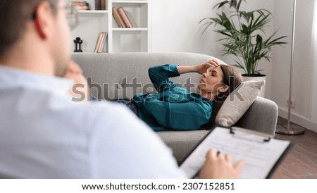 A young woman is lying on a couch at a psychologist's appointment. A brunette male psychologist in glasses conducts a psychological session sitting on a chair in his office. Royalty-Free Stock Photo #2307152851