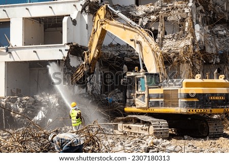 excavator equipped with a hydraulic hammer demolishes old building while a worker sprays water to minimize dust Royalty-Free Stock Photo #2307180313
