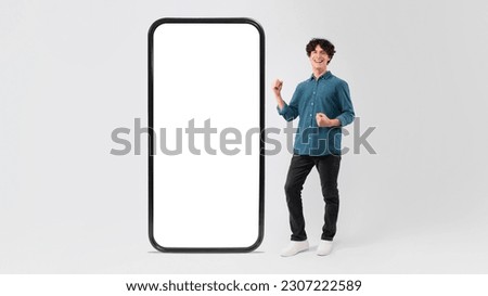 Online Success. Happy Young Man Posing With Big Cellphone Displaying Empty Screen And Gesturing Yes In Joy, Celebrating Great Mobile News Over White Studio Background. Panorama, Mockup