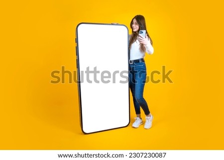Big smartphone, full body length caucasian woman leaning big smartphone. Millennial lady holding mobile phone and leaning huge one with empty blank screen for mockup. Yellow background, copy space. 