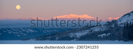 Incredible winter morning sunrise in northern Canada with pink, purple pastel tones covering the snow capped mountain range with frozen lake in foreground. Beautiful moon set in sky above.  Royalty-Free Stock Photo #2307241255