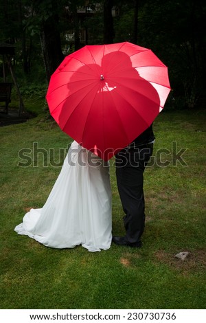 Love concept: Bride and groom kiss behind red heart umbrella, back lit with off camera flash