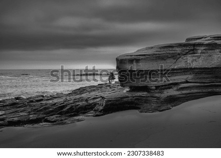 A UNIDENTIFIED PERSON SITTING ON A BUCKET AT THE BEACH FISHING IN THE EARLY MORNIG BY A ROCKY OUT CROPPING IN LA JOLLA CALIFORNIA IN BLACK AND WHITE