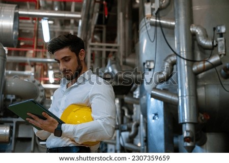 Professional electrical engineer in safety uniform working on digital tablet at factory site. Industrial technician worker maintenance and checking power control system at manufacturing plant room.