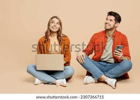 Full body young couple two friends family IT man woman wearing casual clothes together sit hold work on laptop pc computer use mobile cell phone isolated on pastel plain light beige color background