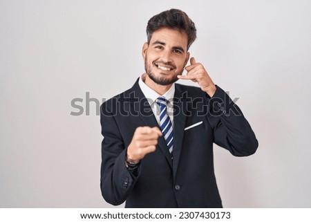 Young hispanic man with tattoos wearing business suit and tie smiling doing talking on the telephone gesture and pointing to you. call me. 