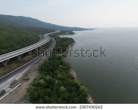 A bird's-eye view of a rest stop indicated by a yellow sign. Along the Lam Ta Klong Dam, Thailand. Cars speeding along the road amidst mountains, rivers and sky.