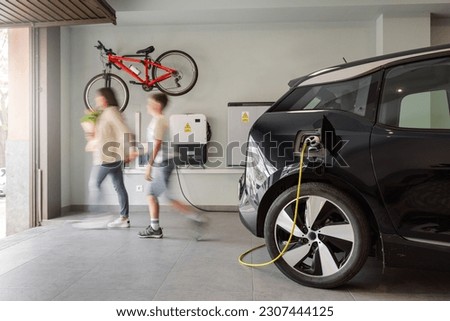 Electric vehicle charging station in a private home with an unrecognizable blurred family leaving the house, with a bicycle hanging on the wall. Copy-space, horizontal., motion blur.