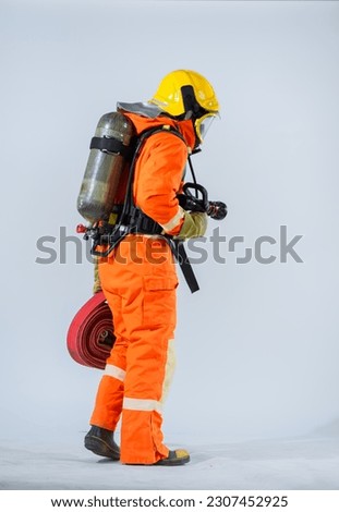 The professional firefighter is standing with his back turned holding a fire hose and wearing an oxygen tank on his back while looking down at the floor on a white background.