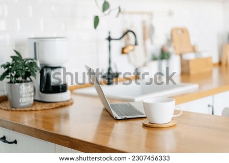 Cup of cofee and laptop on wooden table, view on white kitchen in modern style, drip coffee maker on background