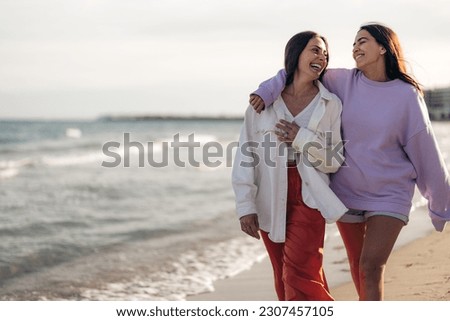 Female friends enjoying the day at sea coast, walking with arms around each other and smiling. Two happy Hispanic girls brunettes in casual clothes spending time by the sea. Royalty-Free Stock Photo #2307457105