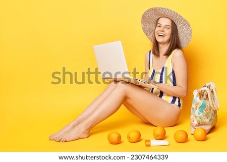 Happy optimistic woman wearing striped swimming suit and hat isolated on yellow background working on notebook laughing happily empty space.