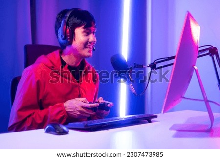 Professional gamer casting podcast walkthrough review. Young asian man sitting on chair with computer pc and microphone. Happy male Streamer wearing headphone playing game online in room neon light Royalty-Free Stock Photo #2307473985