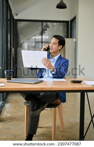 Portrait of a professional millennial Asian businessman looking some details on a paper while talking on the phone with his business partner.