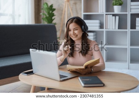 Portrait of a teenage Asian woman using a laptop computer, wearing headphones and using a notebook to study online via video conferencing on a wooden desk in living room resting cozy