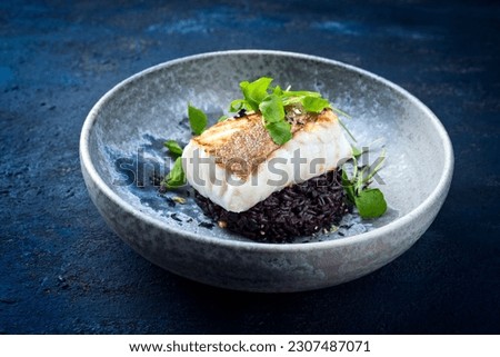 Modern style traditional fried skrei cod fish filet with portulaca lettuce, and black rice served as close-up on ceramic design plate with copy space