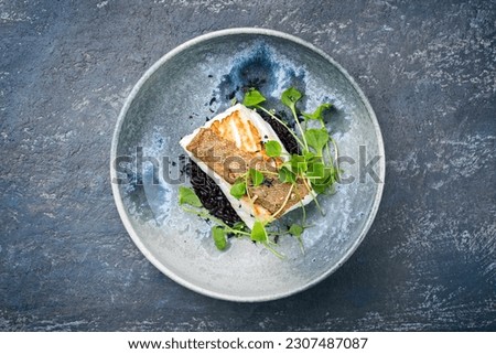 Modern style traditional fried skrei cod fish filet with portulaca lettuce, and black rice served as top view on ceramic design plate with copy space