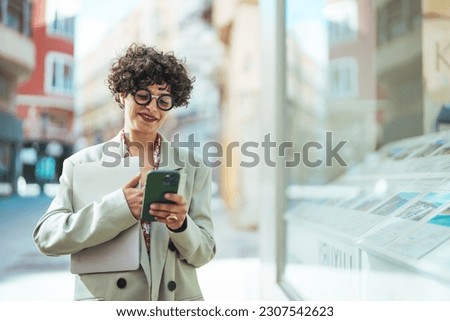 Woman with laptop and smartphone, standing on street of city centre, smiling at camera. Business woman leader wearing suit standing in big city using cell phone platform applications.