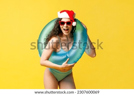 attractive girl in swimsuit and sunglasses in santa hat and with an inflatable swim ring smiles on yellow isolated background for the new year, woman goes to travel on vacation for christmas