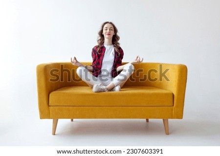 young cute girl sits in lotus position on soft comfortable sofa and meditates, woman does yoga on yellow couch on white isolated background