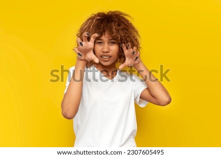 young american curly woman in white t-shirt shows her claws with her fingers and imitates tiger, african girl growls and bares her teeth on pink isolated background Royalty-Free Stock Photo #2307605495
