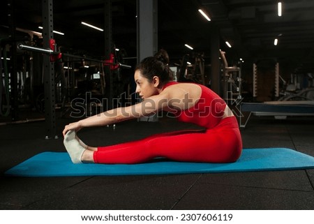 athletic woman in red sportswear sitting on yoga matte in black gym and warming up, girl doing yoga and stretching, attractive woman in fitness club doing flexibility exercise Royalty-Free Stock Photo #2307606119