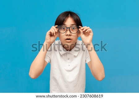 shocked asian boy schoolboy of twelve years old looks into glasses and is surprised on blue isolated background, korean child looks in amazement at the camera and explores and searches
