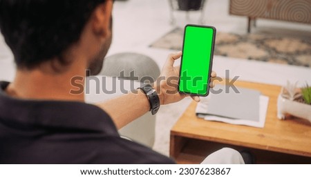 Over the Shoulder Indian Man Using Smartphone with Green Green Screen Screen: Browsing Social Media, Staying Connected with Friends, Doing Online Shopping, Close-up .