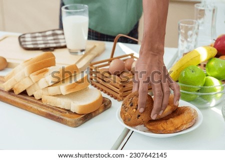 Close-up hand young asian man reaching for cookie placed in plate on kitchen table, grabbed a cookie ate it pick up glass milk before walking out kitchen, In other hand, holds laptop computer work. Royalty-Free Stock Photo #2307642145