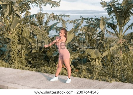 A full body picture of a fit woman wearing a workout outfit and doing arm stretches before jogging. Tall grass and coconut trees in the background