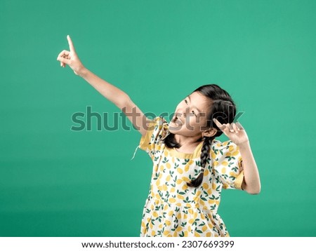 A portrait of a young Asian girl wearing a fruit-patterned dress, seen posing and pointing at something, isolated on a green background.