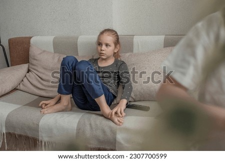 A cute little girl is sitting on the sofa at home in the living room and switches channels on the TV with a remote control. Watching children's shows or videos. Free time after school.