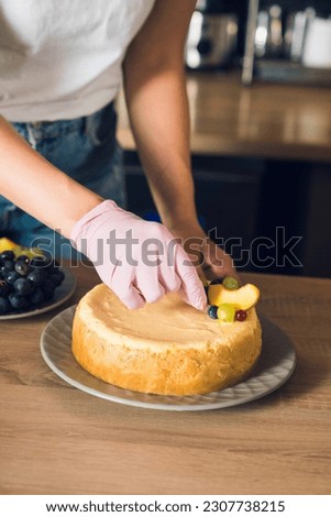 Woman's hands in pink glove decorate top of a cake by fruits