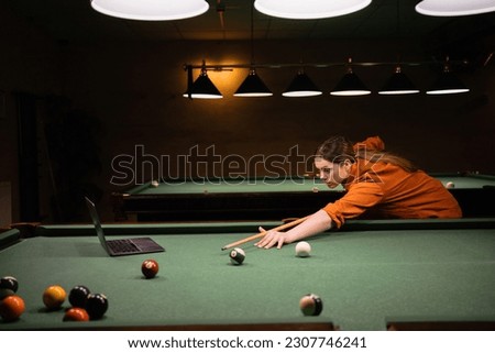 Beautiful girl learning to play billiards watching online video lesson of playing billiards using laptop on pool table Royalty-Free Stock Photo #2307746241