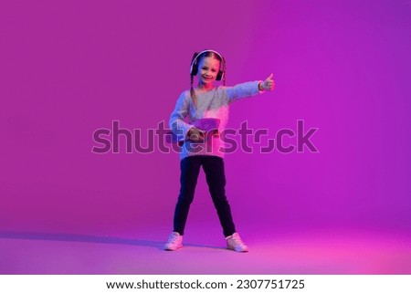 Generation alpha. Happy cheerful cute preteen stylish girl having fun on luminous background, holding skateboard, using wireless headphons, showing thumb up and smiling, copy space
