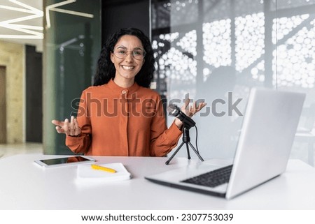 Portrait of young Hispanic woman sitting in office at desk in front of microphone and laptop and recording blog, podcast, webinar. Smiling at the camera.