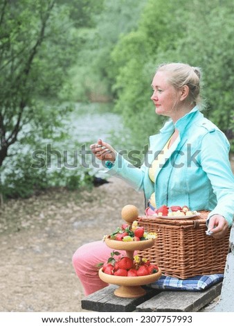 Portrait of a beautiful middle-aged caucasian blonde woman sitting on a bench with a wicker basket and ripe strawberries in a wooden bunk plate in the forest, close-up side view. 