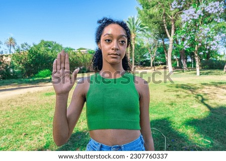 Serious girl asking to stop now, to stop, with raised hand showing palm, with defocused parke background, sunny day with blue sky.