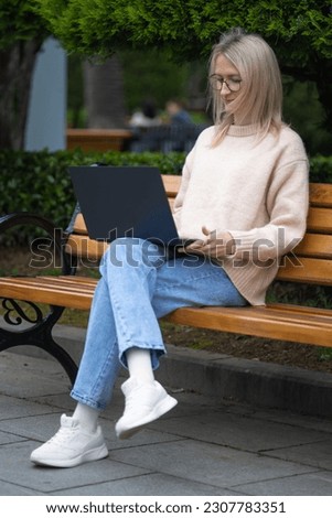 Young smiling blonde woman with glasses is sitting on a park bench with a laptop on her lap. Concept of freelancing, online communication, outdoor learning. Vertical photo