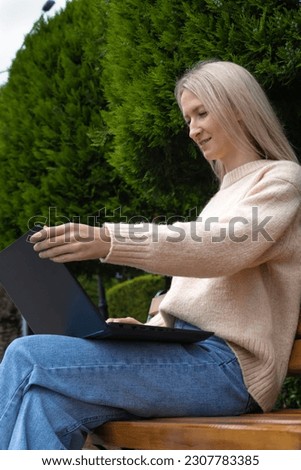 Side view of a young beautiful smiling blonde sitting on a bench with a laptop in the park. Girl opens a laptop in the park sitting on a bench. Vertical photo