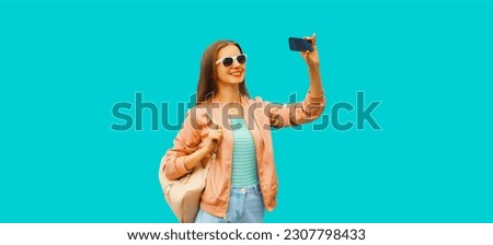 Portrait of happy smiling young woman taking selfie with smartphone wearing backpack on blue background