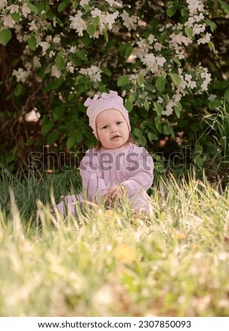 Happy little girl is playing in spring garden. Charming one-year-old girl in pink jumpsuit enjoys walk in fresh air. Child is sitting on green grass. Vertical photography