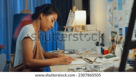 Youth Asia lady wear apron sit front of desk drawing picture on sketchbook create artwork cozy workshop in home at night. Contemporary Painter Abstract Modern Art, Creativity and people concept.