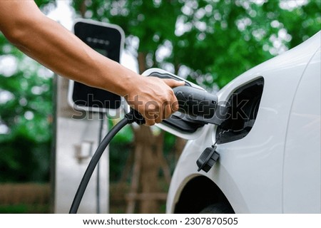 Hand inserting EV charging plug to electric vehicle in focus shot with blurred background of outdoor natural greenery. Progressive sustainable energy powered electric charging station for rechargeable Royalty-Free Stock Photo #2307870505
