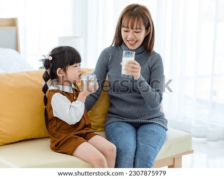 Millennial Asian young pretty female teenager mother nanny babysitter in casual outfit sitting on sofa smiling holding serving delicious milk glass to little cute preschooler daughter girl drinking. Royalty-Free Stock Photo #2307875979