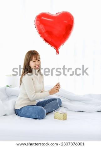Millennial Asian young beautiful female teenager in turtleneck sweater and jeans sitting on bed smiling holding red heart shape helium balloon with surprised paper wrapped ribbon present gift box.