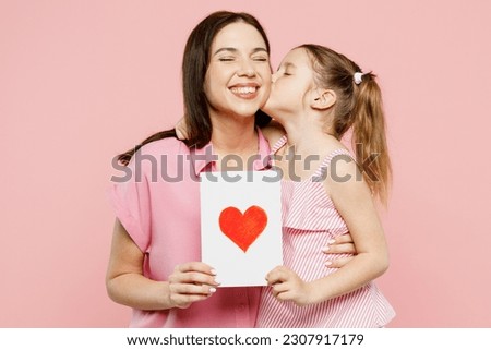 Happy lovely woman wearing casual clothes with child kid girl 6-7 years old. Daughter give mother postcard with heart, hug and kiss isolated on plain pastel pink background. Family parent day concept