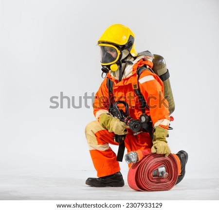 Professional firefighter is sitting turned sideways kneeling down holding a fire hose and wearing an oxygen tank on his back while looking sideways on a white background.